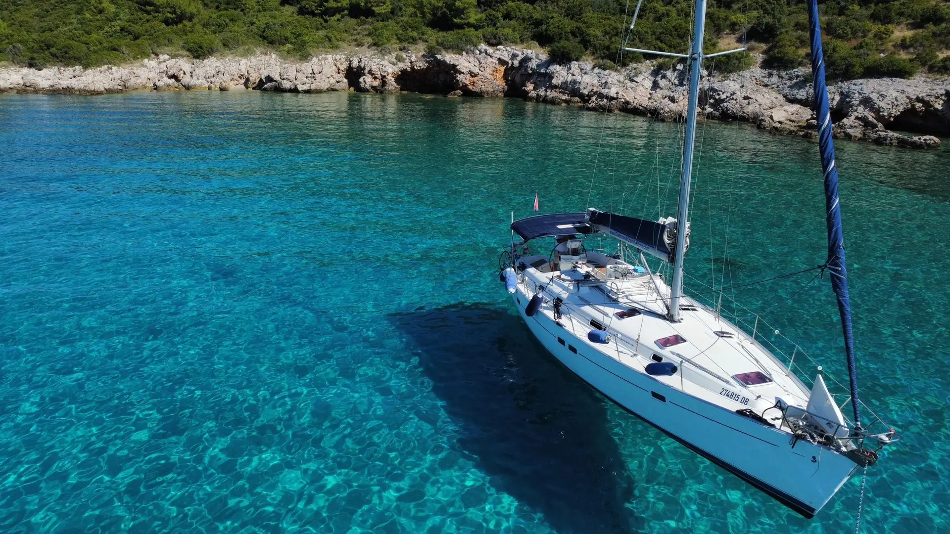 Beneteau Oceanis 411 sailing yacht at anchor in clear turquoise water beside a rocky, pine-covered shore